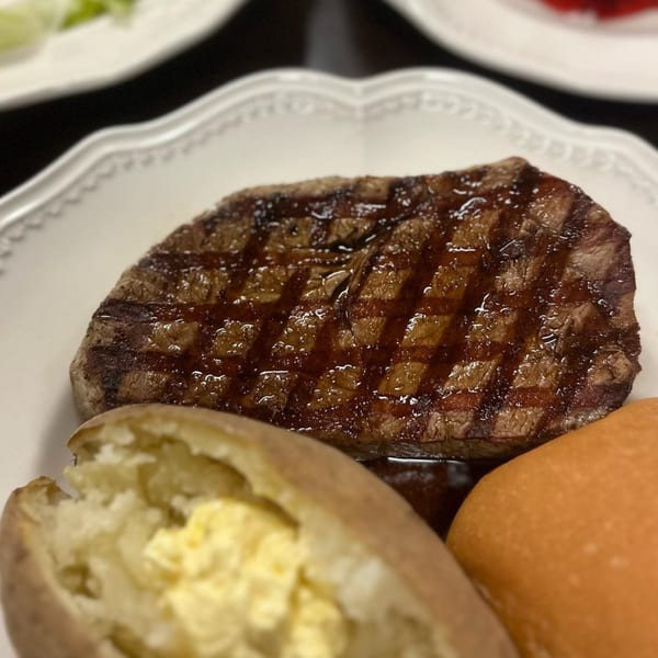 A grilled Steak-Out steak with grill marks, a baked potato with butter, and a dinner roll.